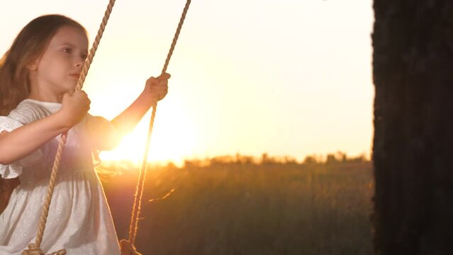 Girl with hair swayed by wind enjoys riding swing with sunset in background. Girl with long hair enjoys swing with sun setting over field. Girl with loose hair glimmering in fading light rides swing