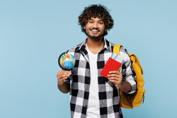 Young Indian student wear shirt casual clothes backpack bag hold globe boarding passport tickets, travel abroad isolated on plain pastel light blue background. High school university college concept.