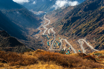 A mesmerizing view of a road passing through a village located deep down in the mountain valley. The beautiful valley is surrounded by dark mountains covered with brown grass and clouds. Sikkim, India