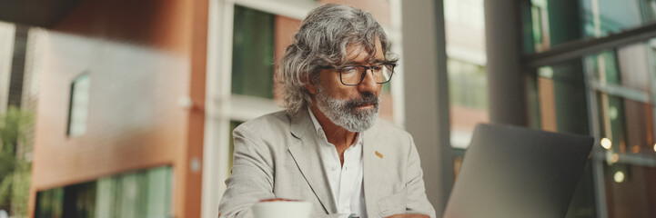 Mature businessman with beard in eyeglasses wearing gray jacket sits on cafe. Panorama of middle aged manager successful man user on laptop pc computer sit at cafe outdoors - Powered by Adobe