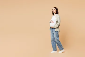 Fototapete Full body side view young minded pregnant woman future mom in grey shirt with belly stomach stroking put hand on tummy with baby isolated on plain beige background Maternity family pregnancy concept © ViDi Studio