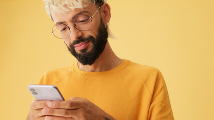 Guy with glasses, dressed in yellow T-shirt, laughs while reading sms, watching video on mobile phone, isolated on yellow background in studio