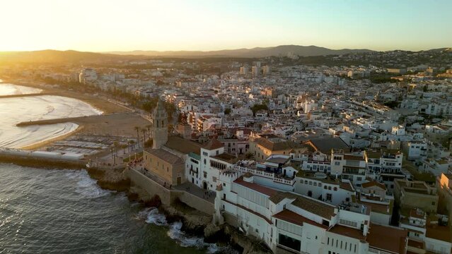 Beautiful orange sunset over Sitges city. Cinematic aerial perspective. drone going forward over the Church of Sant Bartolomeu i Santa Tecla. Golden hour colours. Sun reflecting on the buildings.