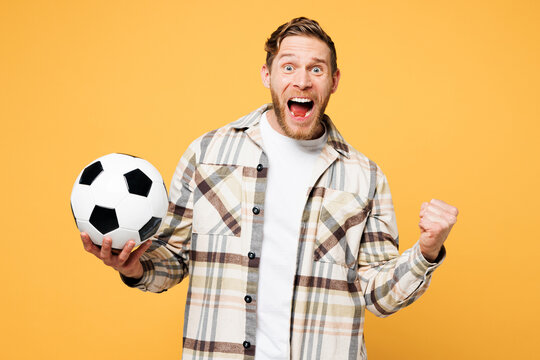 Side view young happy man fan wears brown shirt casual clothes cheer up support football sport team hold in hand soccer ball watch tv live stream do winner gesture isolated on plain yellow background.