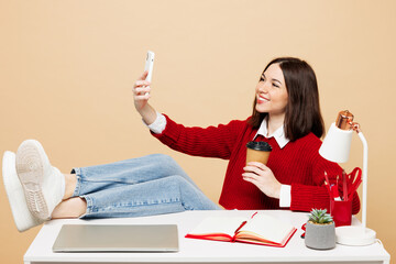 Young employee business woman wear red sweater sit work at office put legs on desk with pc laptop do selfie shot on mobile cell phone drink coffee isolated on plain beige background. Career concept.