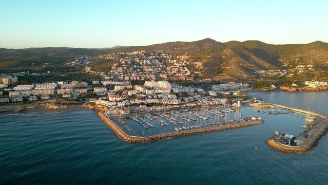 Cinematic aerial view of Sitges Marina at sunset. Drone going forward. Luxury boats and yacht docked in the port. Beautiful warm sunset colours reflecting on the buildings. Famous travel destination.