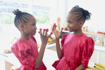 African twin girl sister with curly hair braid African hairstyle eat ripe fresh raspberries from fingers. Happy kid sibling eat fruit and spend time together in kitchen. Cute children in lovely family