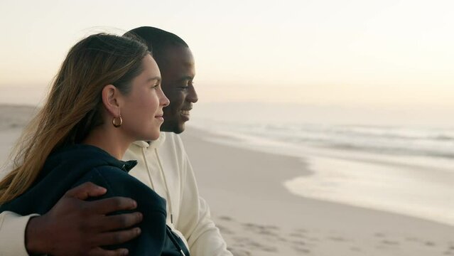 Camera tracks around from front view to rear view of casually dressed loving young couple sitting on sand watching beautiful sunrise morning over beach and sea in South Africa - shot in slow motion