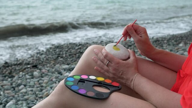 Young woman painting with watercolors on the beach. Hobby and leisure concept. Talent and creativity. Drawing on stones.