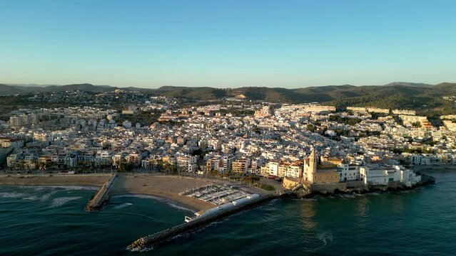 Cinematic aerial view of famouse Sitges city, at sunset. Panoramic view of the city. View of Church of Sant Bartolomeu i Santa Tecla. Waves splashing the cliffs. Beautiful medieval city. Drone forward