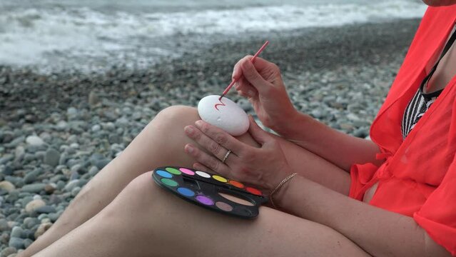 Young woman painting with watercolors on the beach. Hobby and leisure concept. Talent and creativity. Drawing on stones.