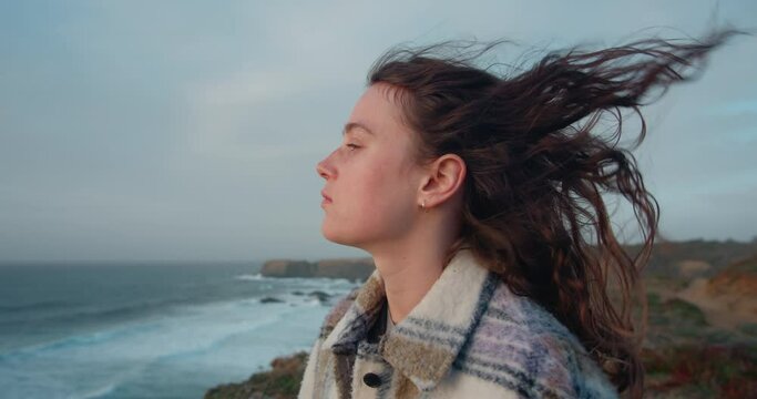 Cinematic shot of young woman standing on a cliff, turning head and smiling into camera. Strong ocean breeze or sea wind blow her hair. Female dreamer breath deeply and feel freedom. 