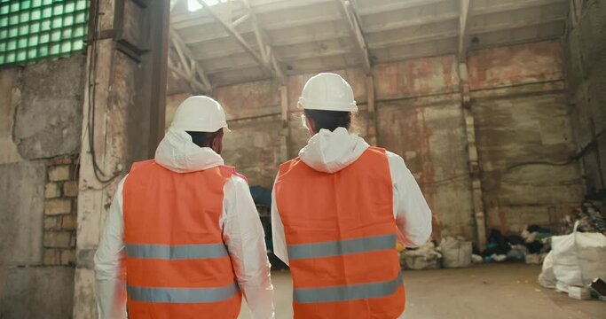 Rear view of two male colleagues in white uniforms and orange vests going out into the premises of a huge waste recycling and sorting plant. A Black man and a brunette man with a beard are talking and