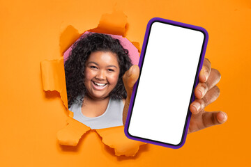 Cheerful young woman with curly hair peering through a torn orange paper, presenting a smartphone...