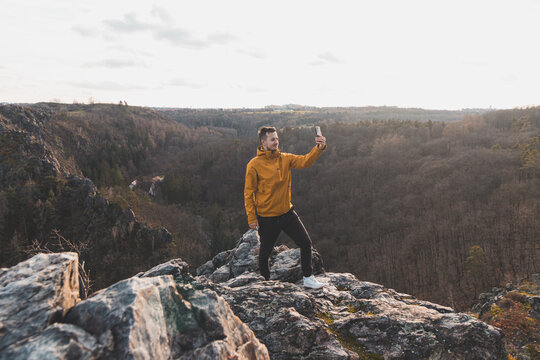 Traveller in a yellow jacket standing on the edge of a cliff taking a selfie on his mobile phone at sunset.  Divoka Sarka valley, Prague