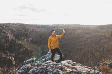 Traveller in a yellow jacket standing on the edge of a cliff taking a selfie on his mobile phone at sunset.  Divoka Sarka valley, Prague