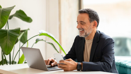 Smiling mature businessman multitasking with a smartphone and laptop in a bright office