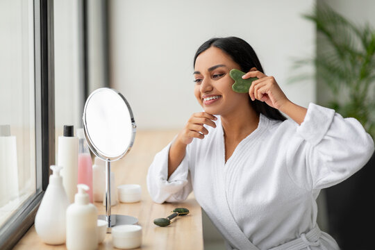 Smiling indian woman using gua sha, looking at mirror
