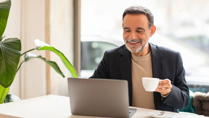 Joyful mature man with a beard working on a laptop and enjoying a cup of coffee in a bright office