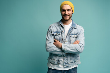 Obraz premium Portrait of stylish brutal young bearded European man wearing yellow hat and jeans jacket, posing at blue blank wall with copy space for your text or promotional content