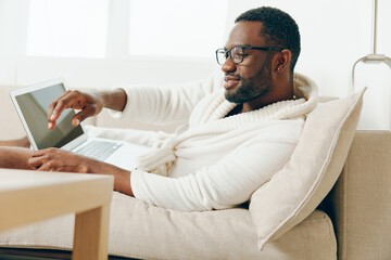 African American freelancer working on a laptop in a modern home The young man dressed in a bathrobe sits on a comfortable sofa in the living room, typing with a smile on his face It's a sunny - obrazy, fototapety, plakaty