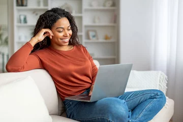 Poster Smiling Black Female Relaxing With Laptop On Couch At Home © Prostock-studio