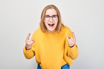 Excited young woman in yellow sweater making rock'n'roll gesture against grey background.