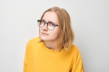 Young person in yellow sweater and glasses looking away thoughtfully against a gray background.