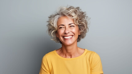 Close up of  mature woman with white skin, grey short hair, wavy hair and a clear yellow t shirt, isolated in a light grey studio. Portrait person. - obrazy, fototapety, plakaty
