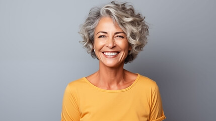 Close up of  mature woman with white skin, grey short hair, wavy hair and a clear yellow t shirt, isolated in a light grey studio. Portrait person. - obrazy, fototapety, plakaty