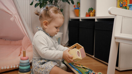 Baby girl playing with toys. He is in the children's room and collects cubes on the floor. An interested baby. In the background is a shelf with toys and a children's tent