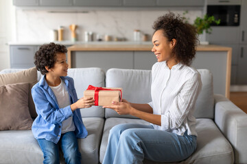 Happy black mother gifting present to her excited son on couch
