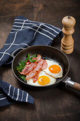 Fried eggs in a frying pan with bacon. Dark background. Salt and pepper shaker on the table.