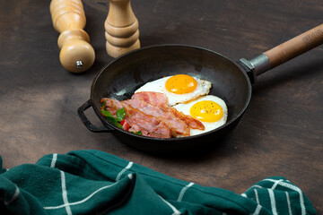 Fried eggs in a frying pan with bacon. Dark background. Salt and pepper shaker on the table.