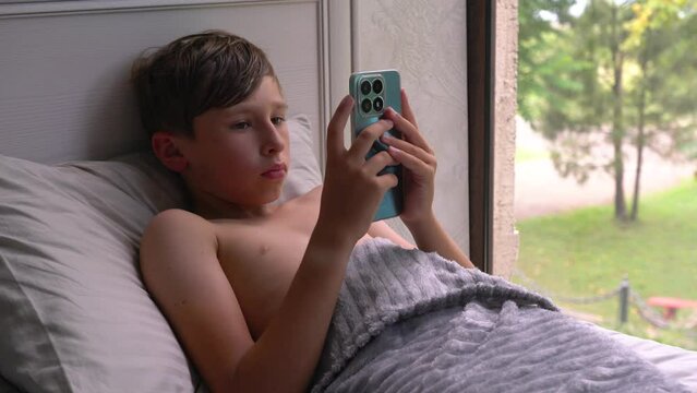 A boy lies on the sofa, scrolling through his phone against the background of the window. Home interior. Teen lifestyle concept