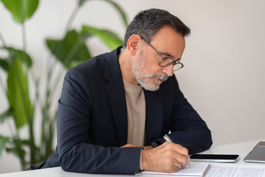 A professional middle-aged man with glasses focused on signing documents at his modern office desk - Powered by Adobe