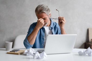 Frustrated middle-aged man massaging his nose and keeping eyes closed while sitting at his working...