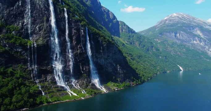 Breathtaking aerial footage of the Seven Sisters waterfall in Norway's Geirangerfjord. A vibrant rainbow arcs over the cascading waters on a sunny day, showcasing the stunning natural beauty. Travel 
