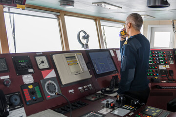 Officer on watch with radio on the navigational bridge. Caucasian man in blue uniform sweater on the bridge of cargo ship.