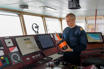 Officer on watch with EPIRB on the navigational bridge. Caucasian man in blue uniform sweater using emergency position indicating radio beacon on the bridge of cargo ship. - obrazy, fototapety, plakaty