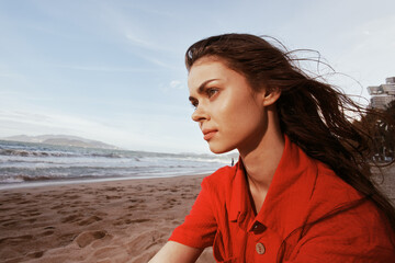 Serene Sunset: Portrait of a Carefree Woman, Enjoying Vacation by the Ocean, Sitting on the Sand, in Fashionable Red Clothing, against a Beautiful Beach Background.