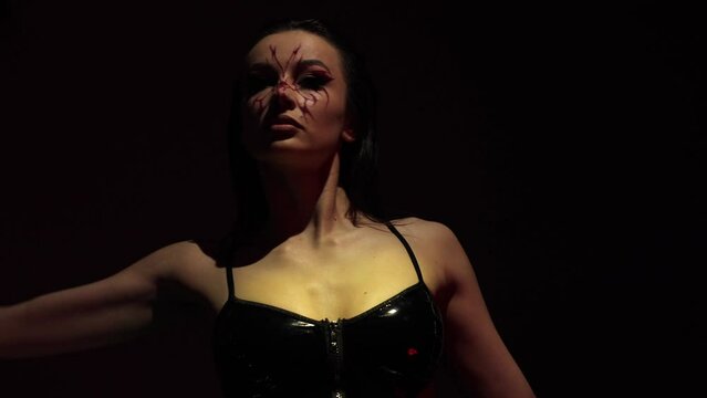 Dancing in the dark, a beautiful girl on a black background. Dance choreography of a young woman, she is dressed in black clothes. Sexy scene, she has weird makeup on her face. Great flexibility