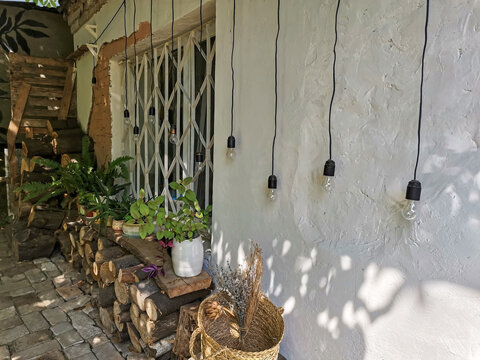 Small cozy summer yard. Logs are stacked along the wall. A variety of potted plants growing under the window. Light bulbs hanging on the wall of the house