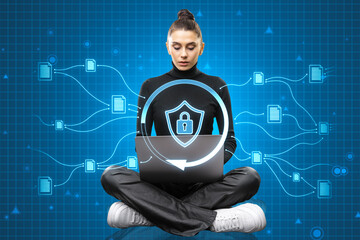 Casual young woman sitting on floor and using laptop with glowing padlock shield hologram on blurry...