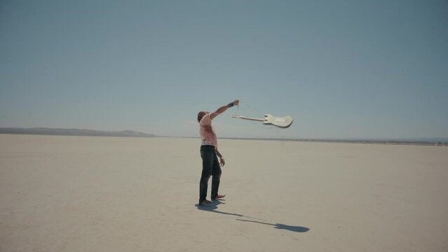 Angry man with red hair throws away guitar with chains. Barefoot and bloody musician in desert. Slow motion acting at dry lake. Feelings of desperate frustration and anger. Warm color correction, 4k
