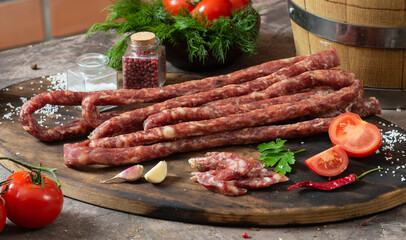 Rustic dry thin sausages among vegetables and herbs on a dark table