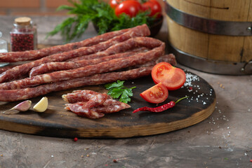 Rustic dry thin sausages among vegetables and herbs on a dark table