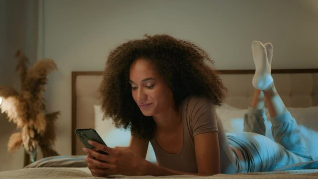 African American ethnic woman 20s girl curly hair scrolling mobile phone relaxing at home evening night insomnia smiling dreaming female lying on bed with smartphone gadget chatting online dating app