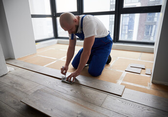 Man in work overalls using metal construction ruler and pen while drawing line on laminate flooring board. Male floor installer preparing laminate plank for installation in apartment under renovation.