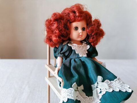 A vintage doll with red hair in a green dress sits on a wooden chair